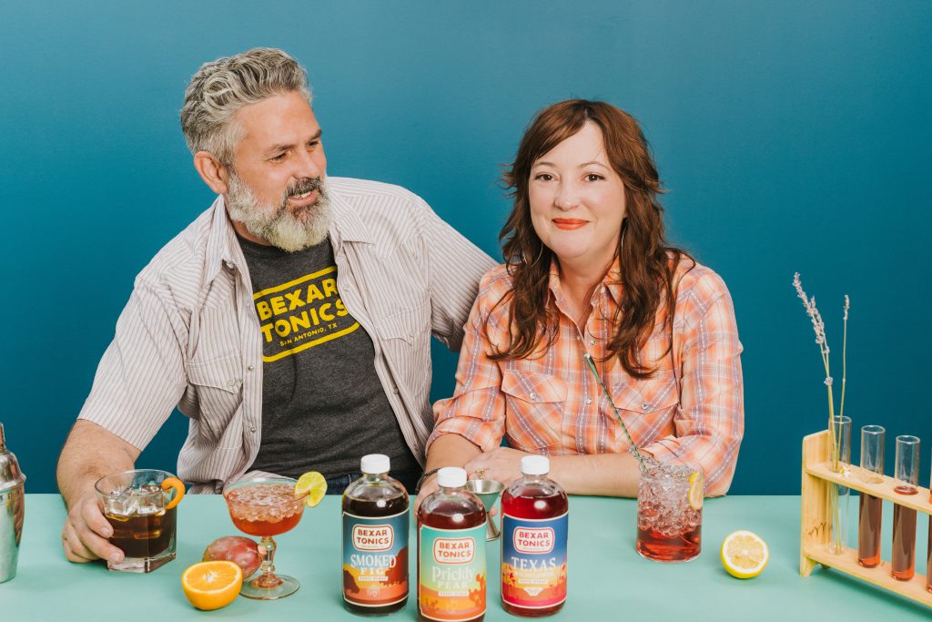 the two creators of bexar tonics sitting at a table with drinks and ingredients